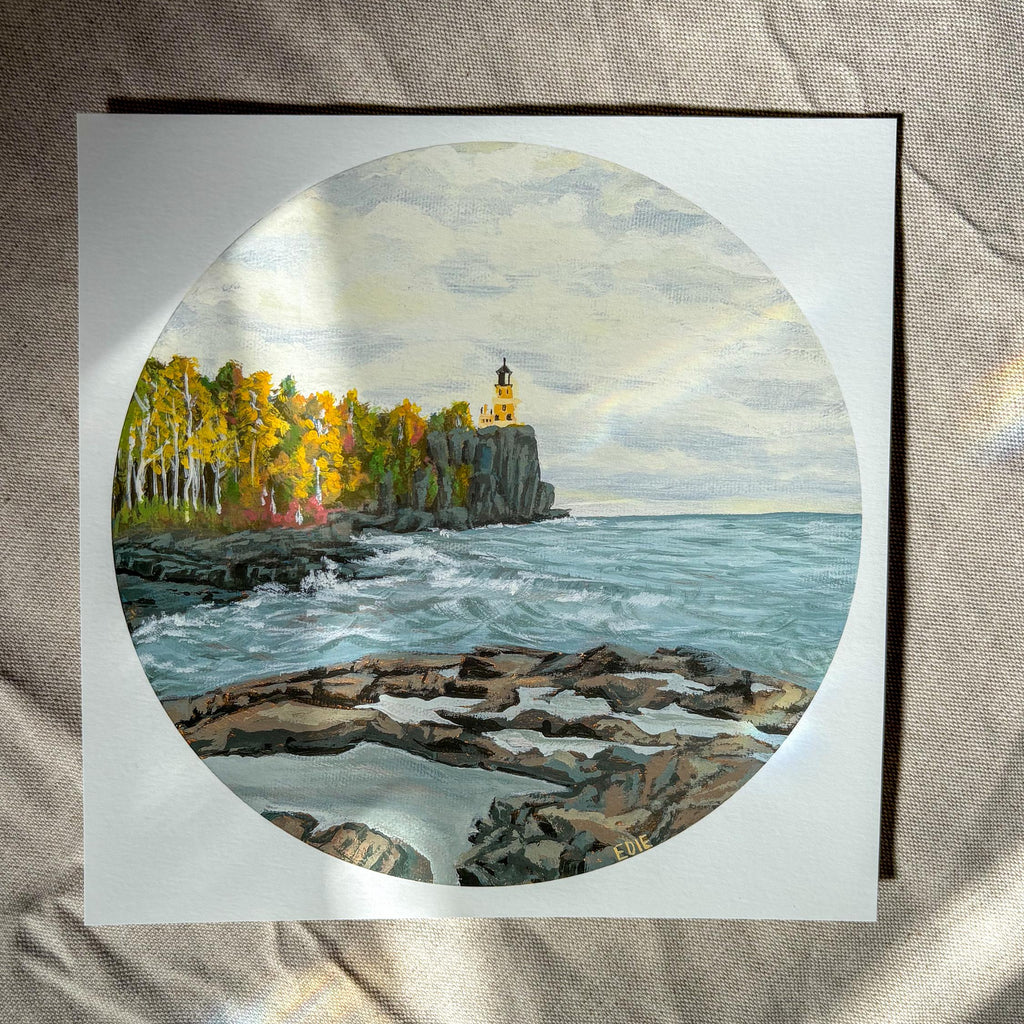 A square art print featuring a circular landscape painting of a rocky coastline with a lighthouse, autumn trees, and blue-gray water, displayed on a beige fabric surface.