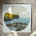 A square art print featuring a circular landscape painting of a rocky coastline with a lighthouse, autumn trees, and blue-gray water, displayed on a beige fabric surface.