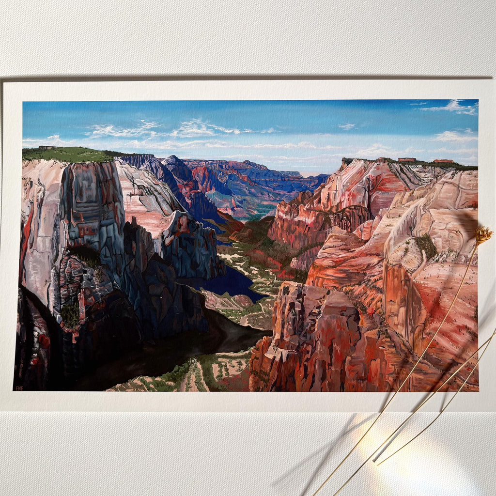 A landscape art print depicting a colorful canyon with red, orange, and blue rock formations under a blue sky, displayed on a white wall with dried flower stems in the foreground.