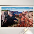 A landscape art print depicting a colorful canyon with red, orange, and blue rock formations under a blue sky, displayed on a white wall with dried flower stems in the foreground.