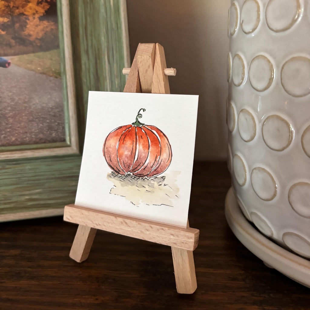 A small square watercolor featuring a pumpkin illustration, displayed on a miniature wooden easel. The easel is set on a dark wooden table next to a large white textured ceramic pot and a framed picture.