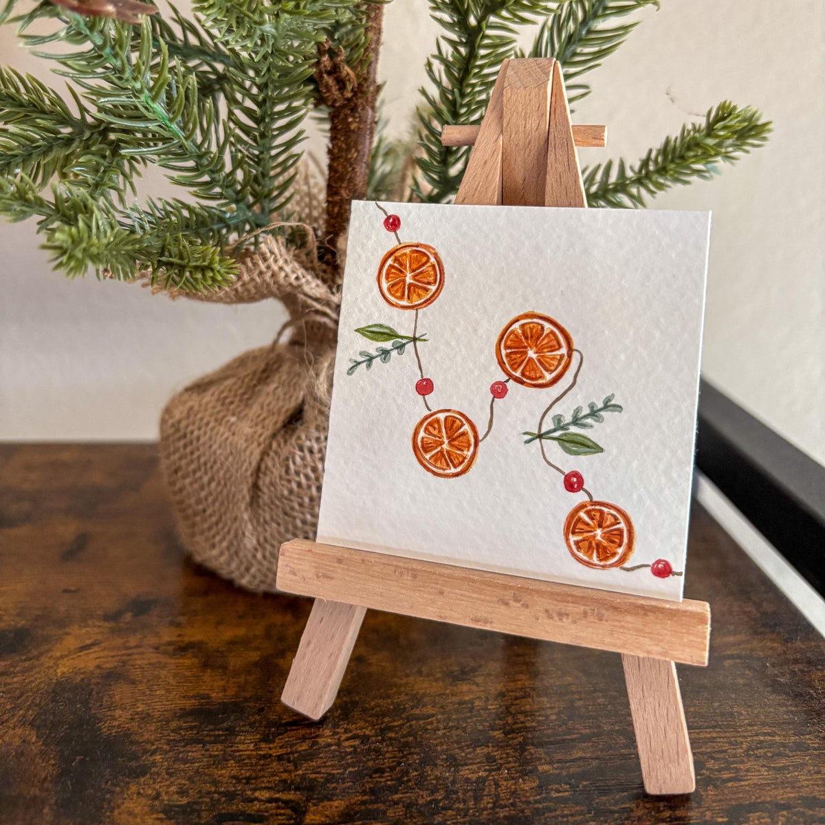 A small square watercolor with an orange slice and berry pattern displayed on a miniature wooden easel, set on a dark wooden table with a potted pine branch in the background.
