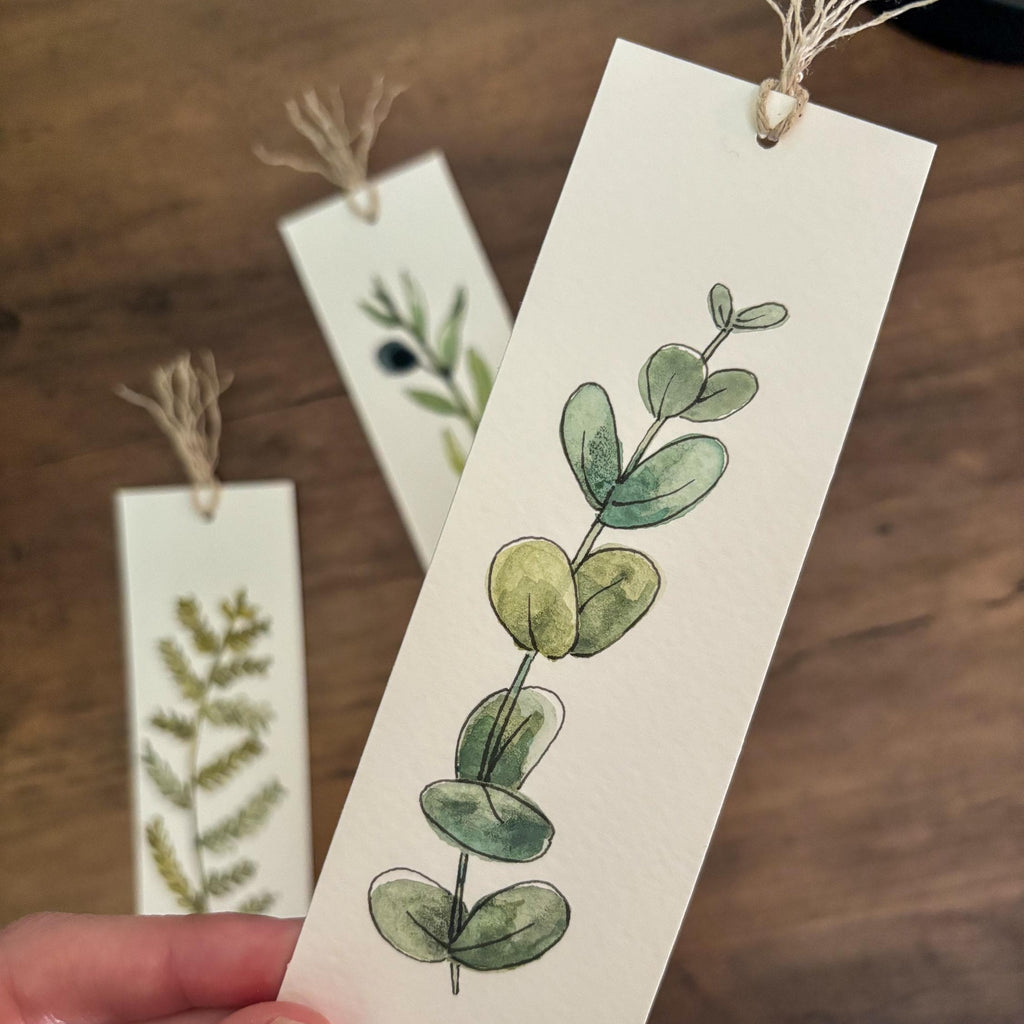 A hand holds a cream-colored paper bookmark featuring a watercolor illustration of green eucalyptus leaves, with two more bookmarks in the background on a wooden surface.