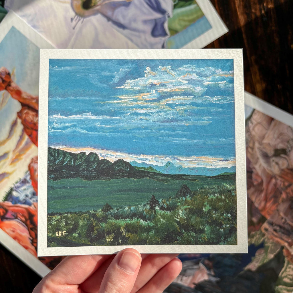 A hand holds a small square landscape painting print with a white border, showing a blue sky, clouds, green fields, and dark mountains. Other colorful art prints are scattered in the background on a dark wooden surface.