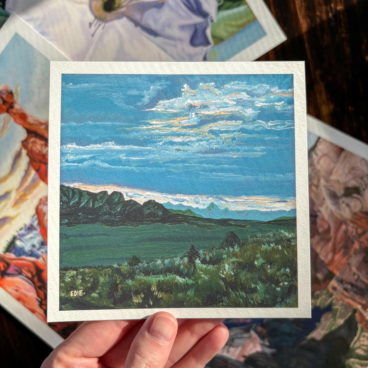 A hand holds a small square landscape painting print with a white border, showing a blue sky, clouds, green fields, and dark mountains. Other colorful art prints are scattered in the background on a dark wooden surface.