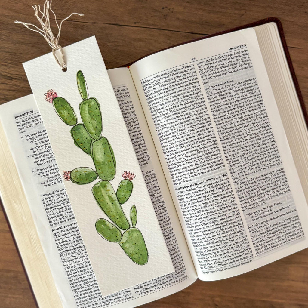 A handmade book bookmark featuring a watercolor illustration of a green prickly pear cactus with small pink flowers, tied to an open book on a wooden table.