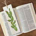 A handmade book bookmark featuring a watercolor illustration of a green prickly pear cactus with small pink flowers, tied to an open book on a wooden table.