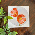 A square art print featuring a circular watercolor illustration of two orange-red  Poppy flowers with yellow centers on a pale blue-gray background, placed on a dark brown wooden surface with green leaves in the foreground.