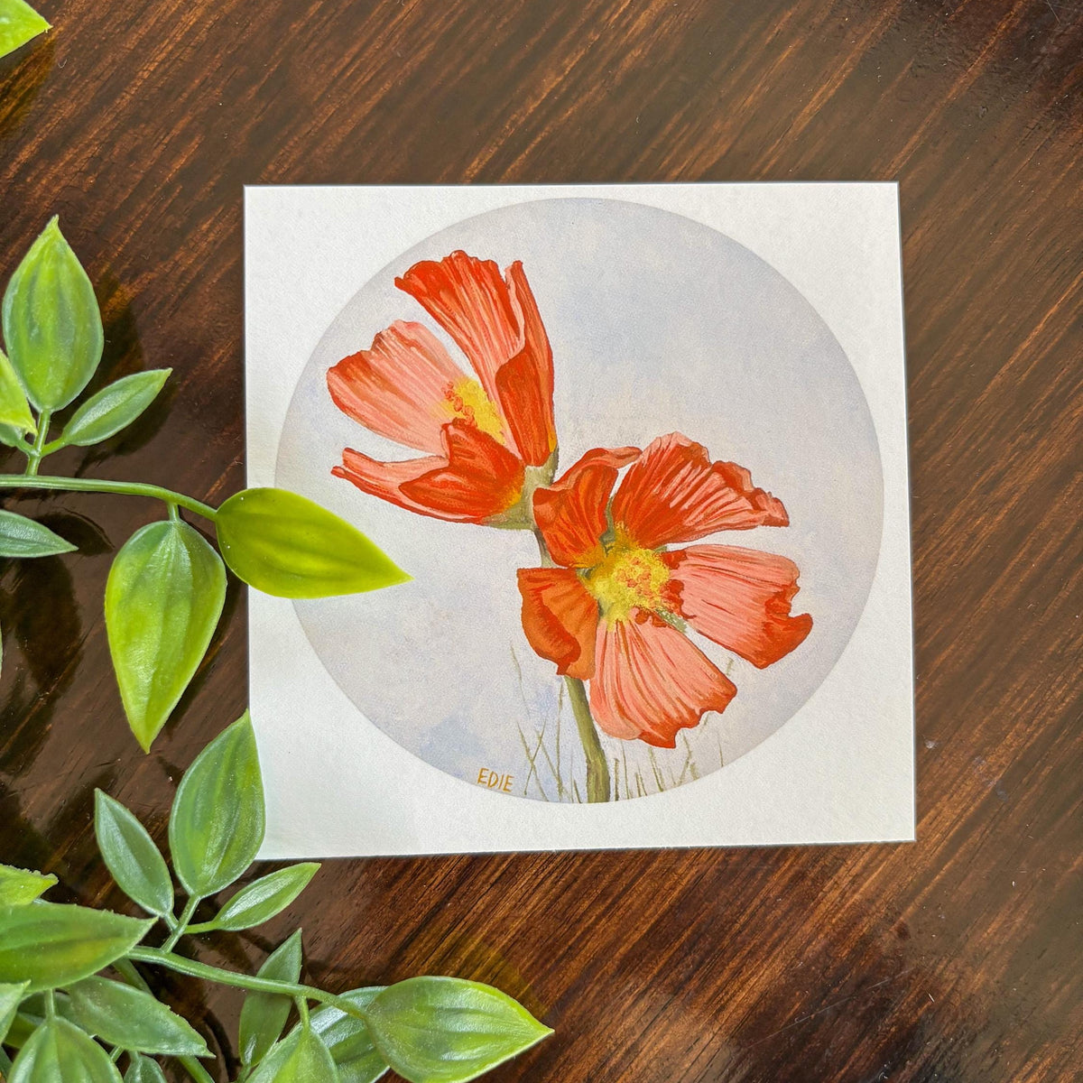 A square art print featuring a circular watercolor illustration of two orange-red  Poppy flowers with yellow centers on a pale blue-gray background, placed on a dark brown wooden surface with green leaves in the foreground.