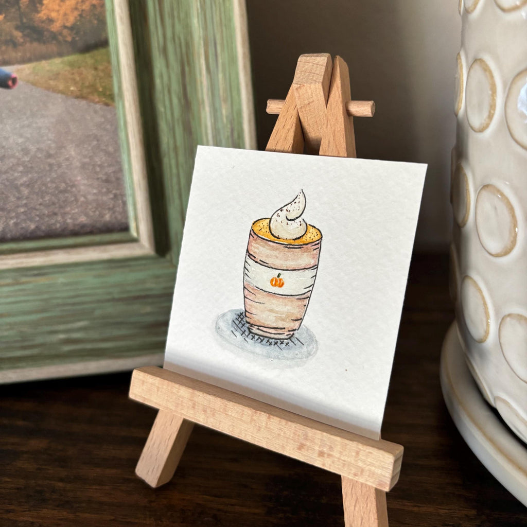 A small watercolor painting of a layered dessert on a white card, displayed on a miniature wooden easel. The scene is styled on a dark wooden table next to a green picture frame and a white ceramic container.