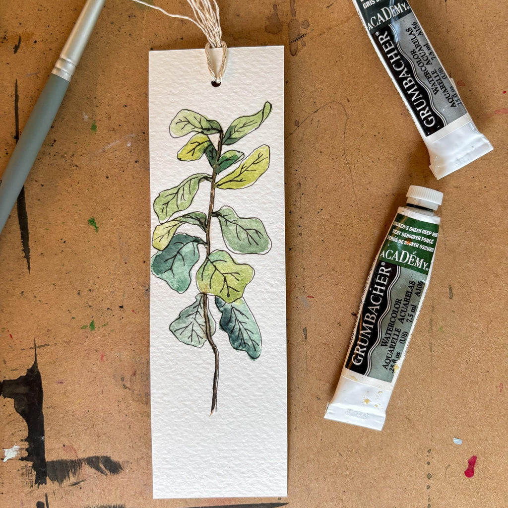 A bookmark with watercolor painting of a green leafy branch, shown as a flat-lay on a brown textured surface with a paintbrush, two tubes of Grumbacher Academi watercolor paint, and a piece of string.