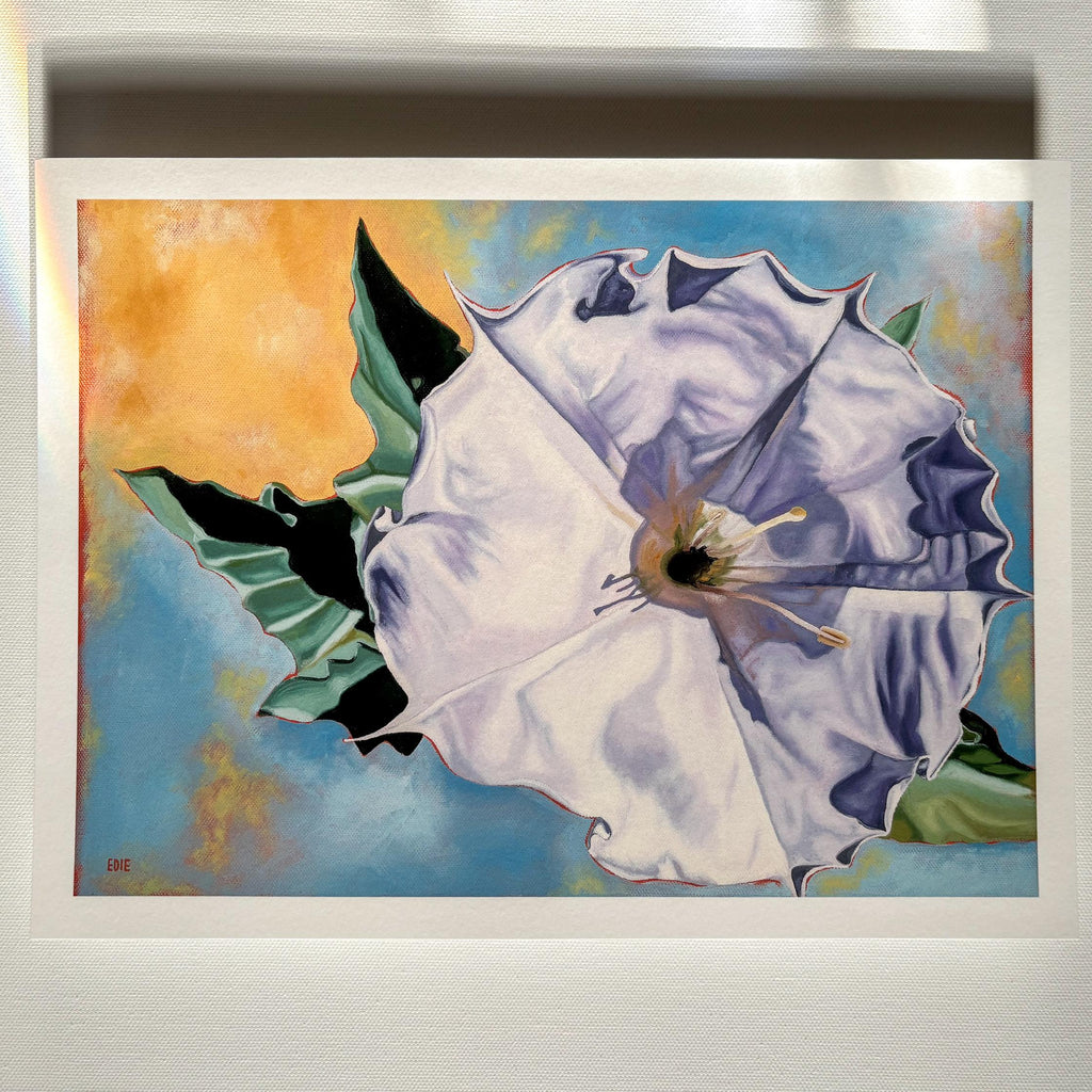 A framed floral art print featuring a large white flower with blue shading, green leaves, and an orange-yellow background, displayed against a light gray wall.