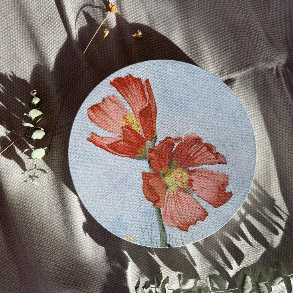 A circular original art piece featuring two red-orange Poppies on a pale blue background, styled on draped gray fabric with leafy props and shadows.