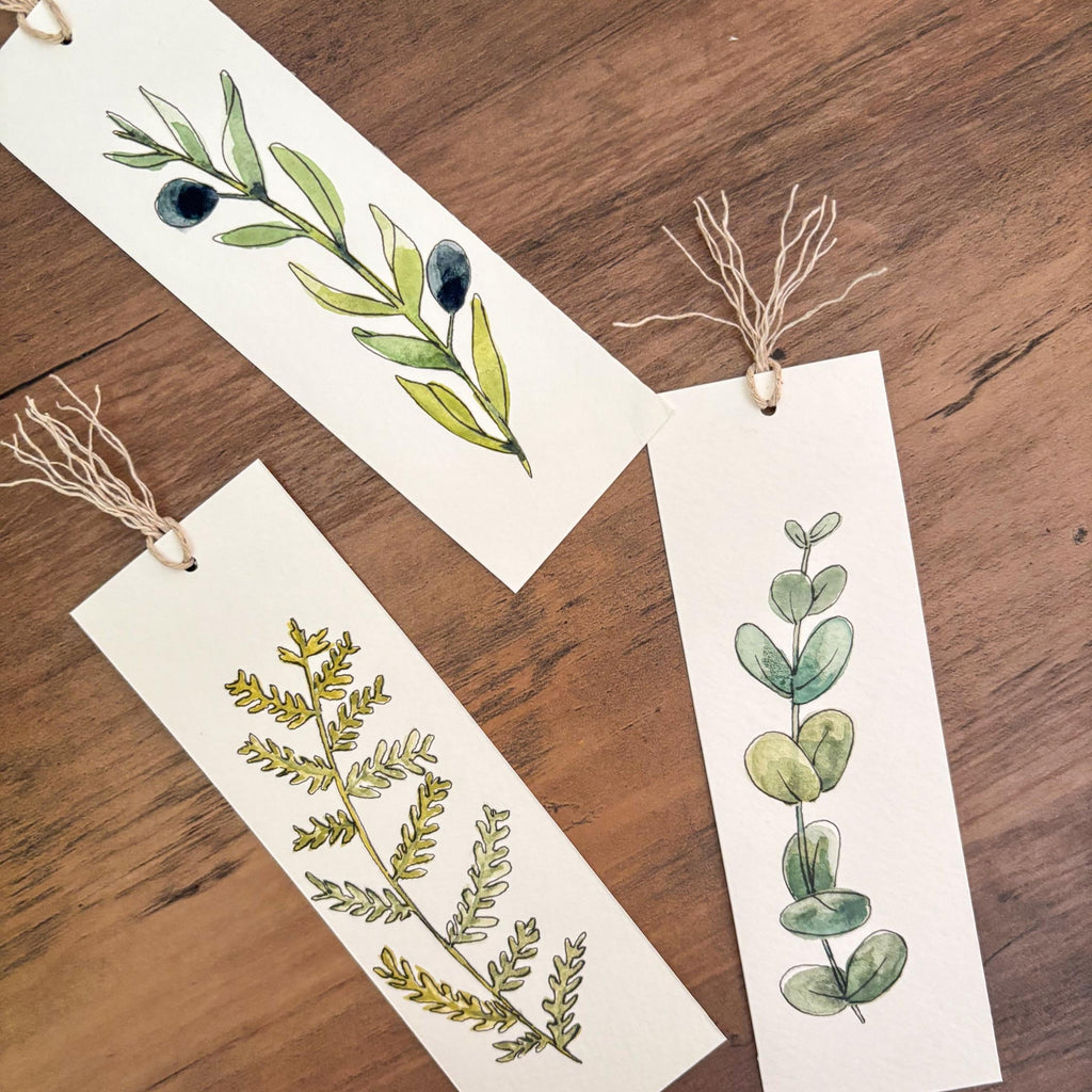 Three rectangular white paper bookmarks with hanging twine are arranged on a wooden surface. Each bookmark features a different watercolor botanical illustration: a green olive branch with dark olives, a yellow-green leafy sprig, and a green eucalyptus stem.