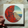 A hand holds a square art print featuring a circular illustration of a canyon landscape with vivid red rock on the left and layered beige cliffs under a blue sky on the right, set against a marble-like surface.