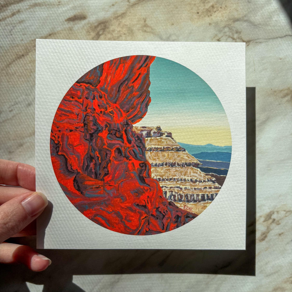 A hand holds a square art print featuring a circular illustration of a canyon landscape with vivid red rock on the left and layered beige cliffs under a blue sky on the right, set against a marble-like surface.