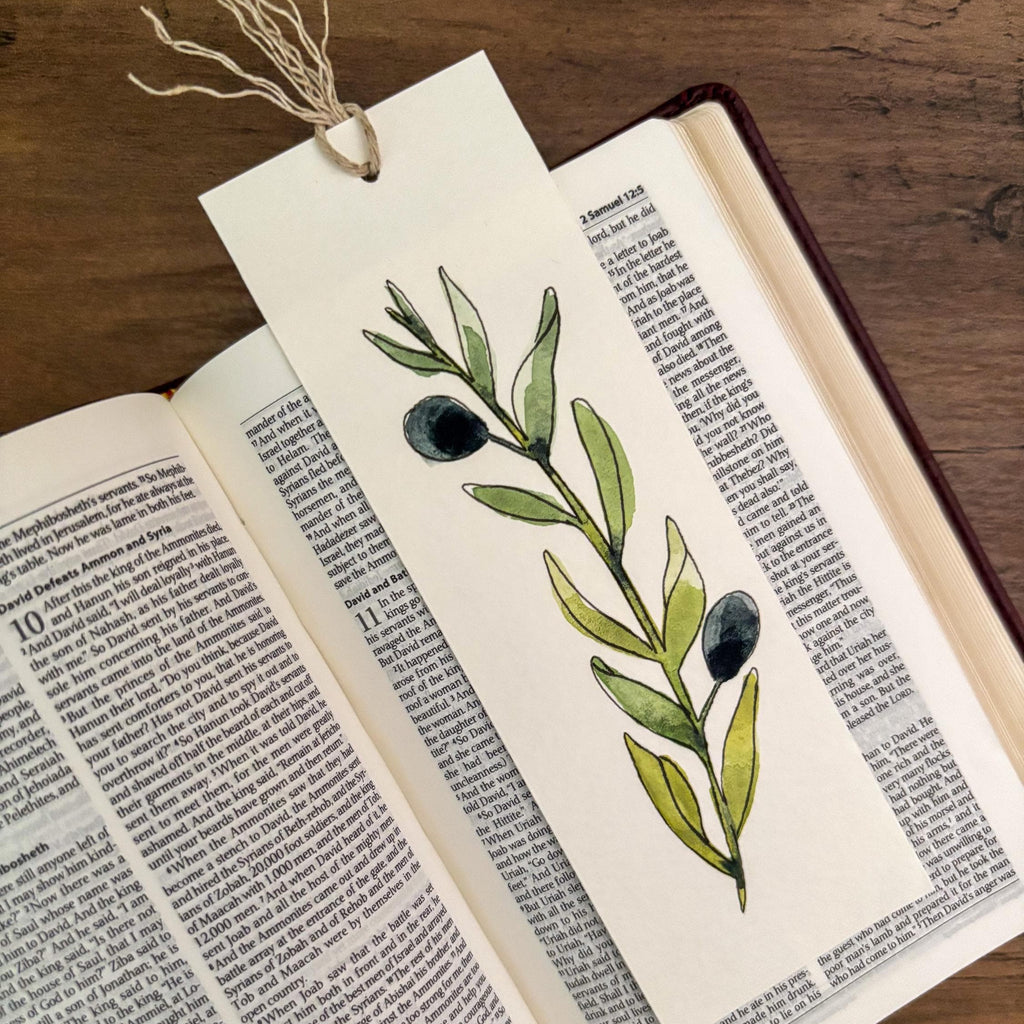 A bookmark with original watercolor olive branch with green leaves and dark olives, tied with a string to an open book on a wooden surface.