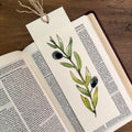A bookmark with original watercolor olive branch with green leaves and dark olives, tied with a string to an open book on a wooden surface.