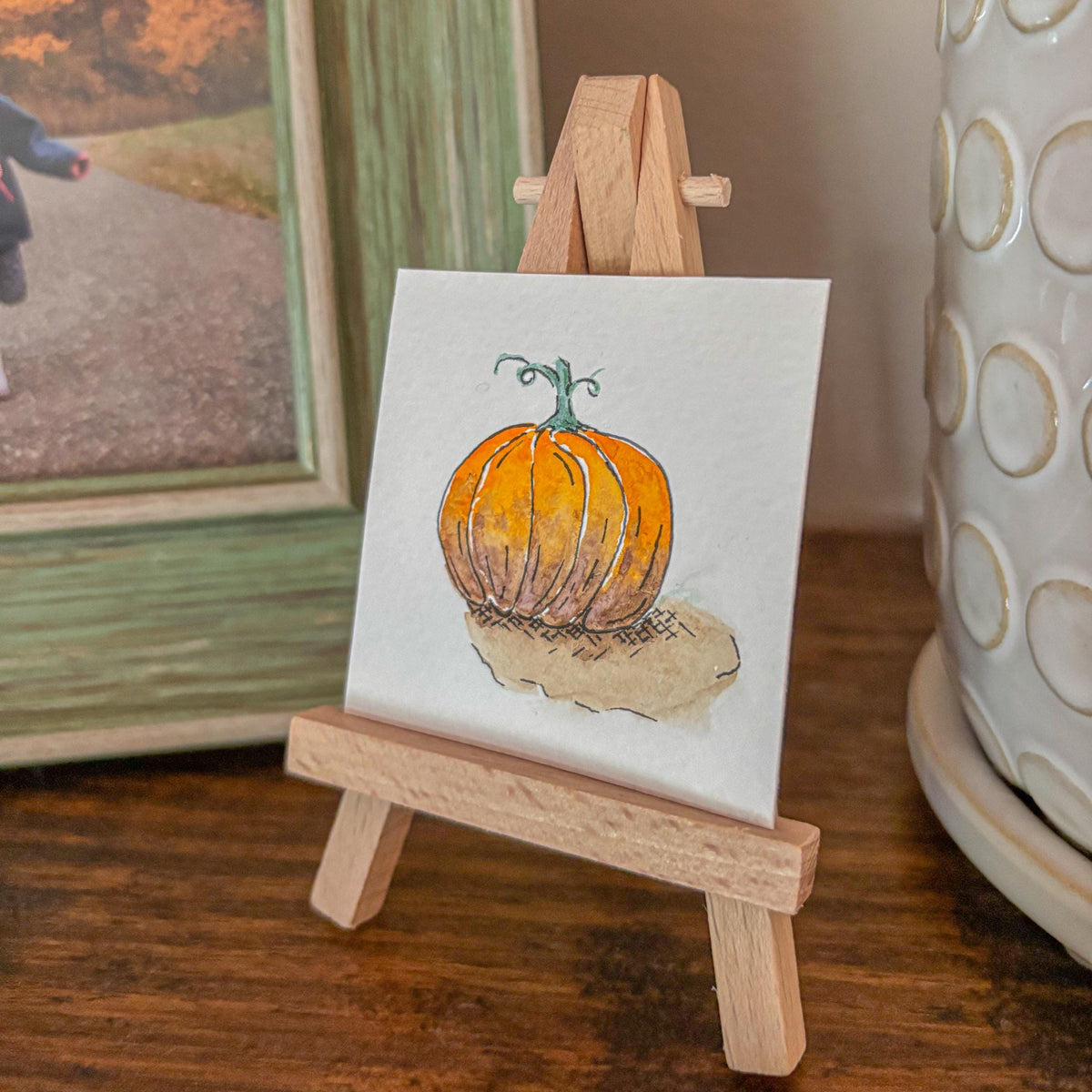 A small hand-painted canvas featuring an orange pumpkin on a white background, displayed on a miniature wooden easel. The scene is styled on a wooden table with a framed autumn photo and a white ceramic pot in the background.