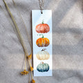 A vertical rectangular paper bookmark with a watercolor illustration of four pumpkins in orange, yellow, cream, and blue-gray tones, hanging from a string on a textured gray fabric background with dried yellow flowers beside it.