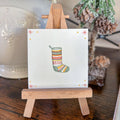 A small square white watercolor art with  a colorful knit Christmas stocking illustration, displayed on a miniature wooden easel stand on a wooden table. A white ceramic pitcher and a frosted decorative tree are blurred in the background.