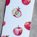 A close-up of a white watercolor bookmark featuring a repeating pattern of red apples, some whole and some sliced open, placed on a textured gray fabric background.