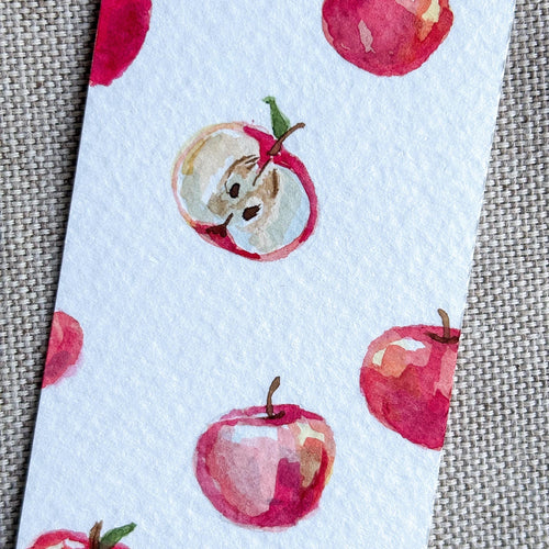 A close-up of a white watercolor bookmark featuring a repeating pattern of red apples, some whole and some sliced open, placed on a textured gray fabric background.