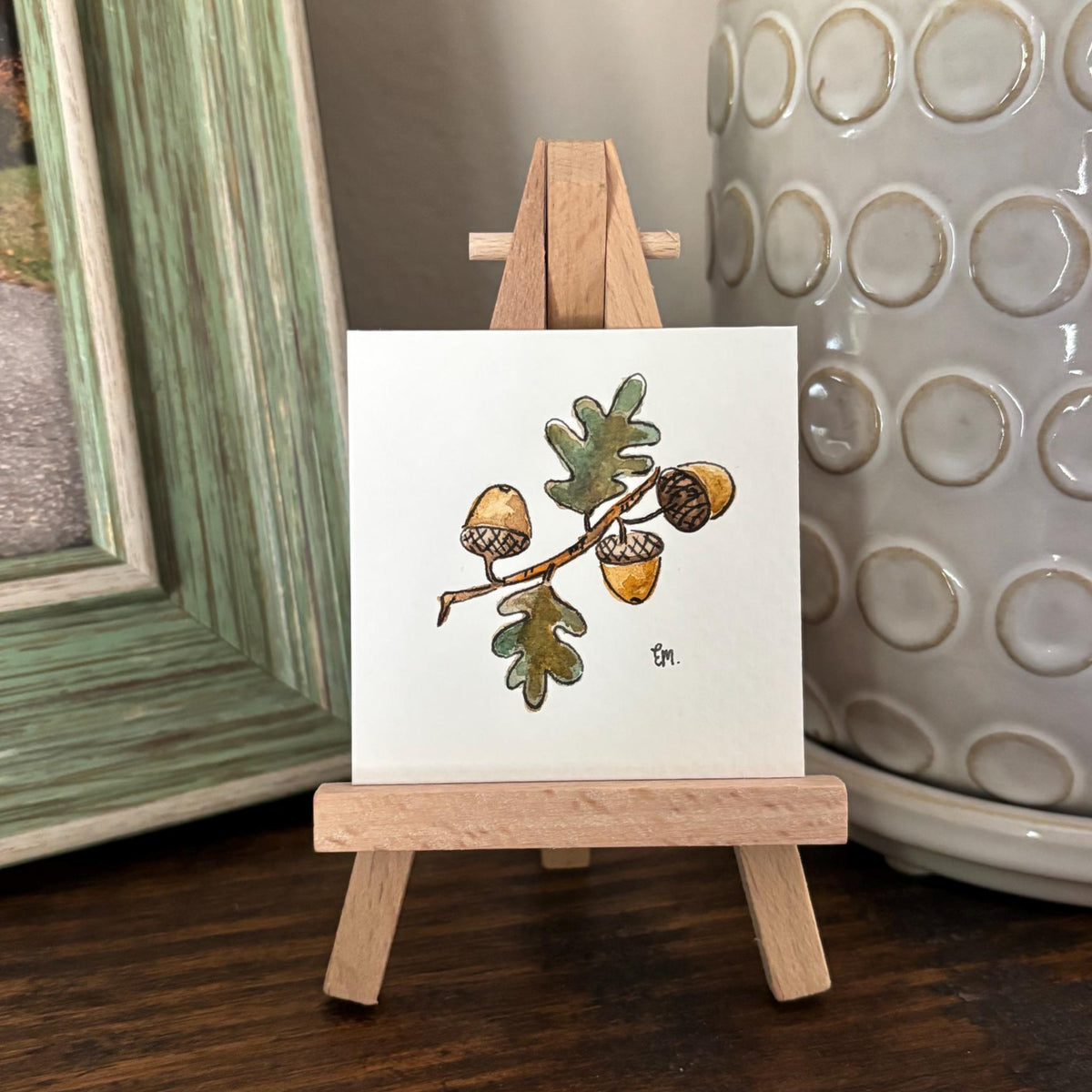 A small watercolor featuring a painted branch with acorns and green leaves, displayed on a miniature wooden easel. The easel sits on a dark wooden table, styled with a green-framed picture and a potted green plant in the background.