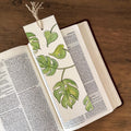 A book bookmark featuring a watercolor illustration of green monstera leaves on a cream page, shown resting on an open book on a wooden table.