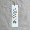 A long rectangular white paper bookmark with a hand-painted botanical design of green and orange leaves and small mushrooms, hanging from a twine string on a textured gray fabric background.
