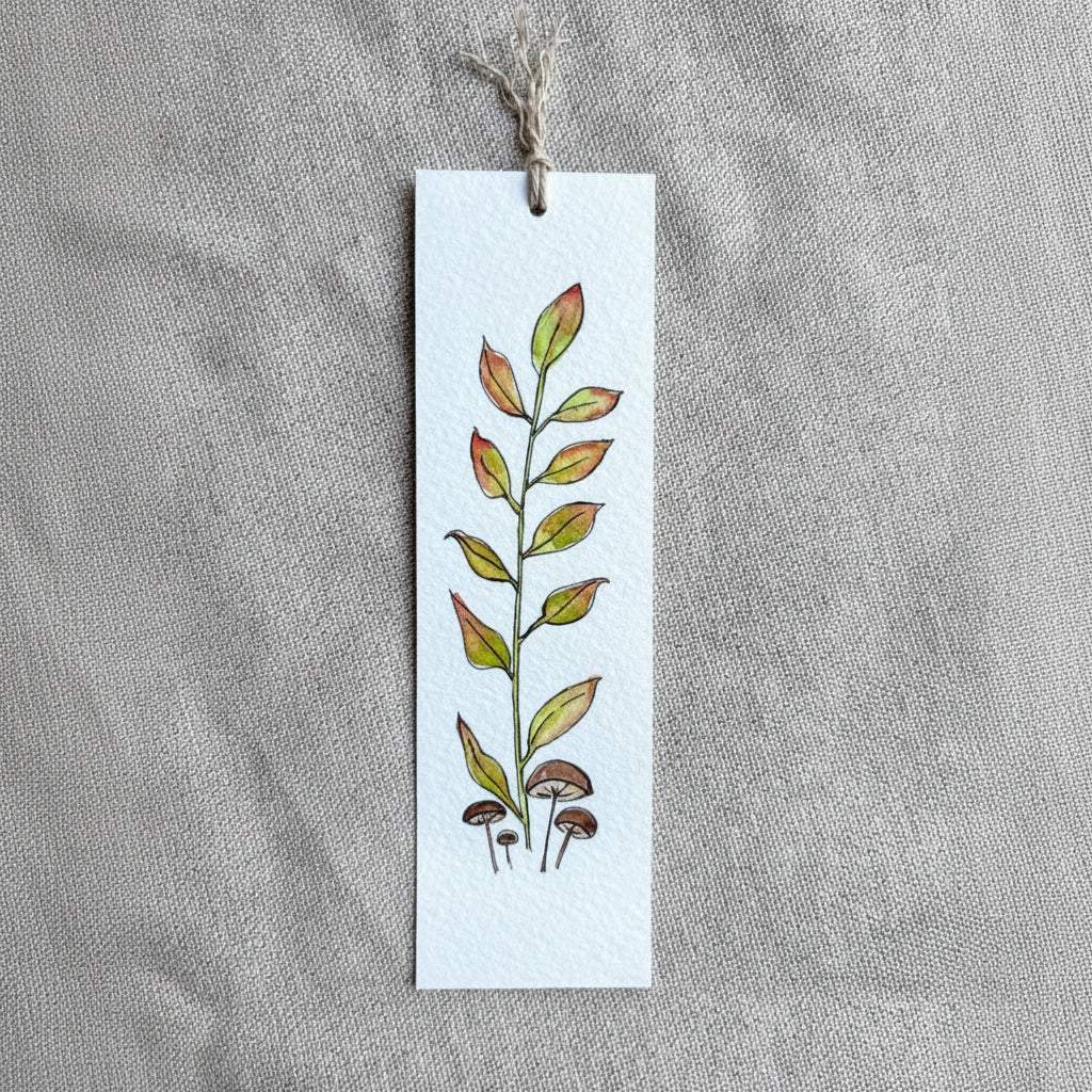 A long rectangular white paper bookmark with a hand-painted botanical design of green and orange leaves and small mushrooms, hanging from a twine string on a textured gray fabric background.